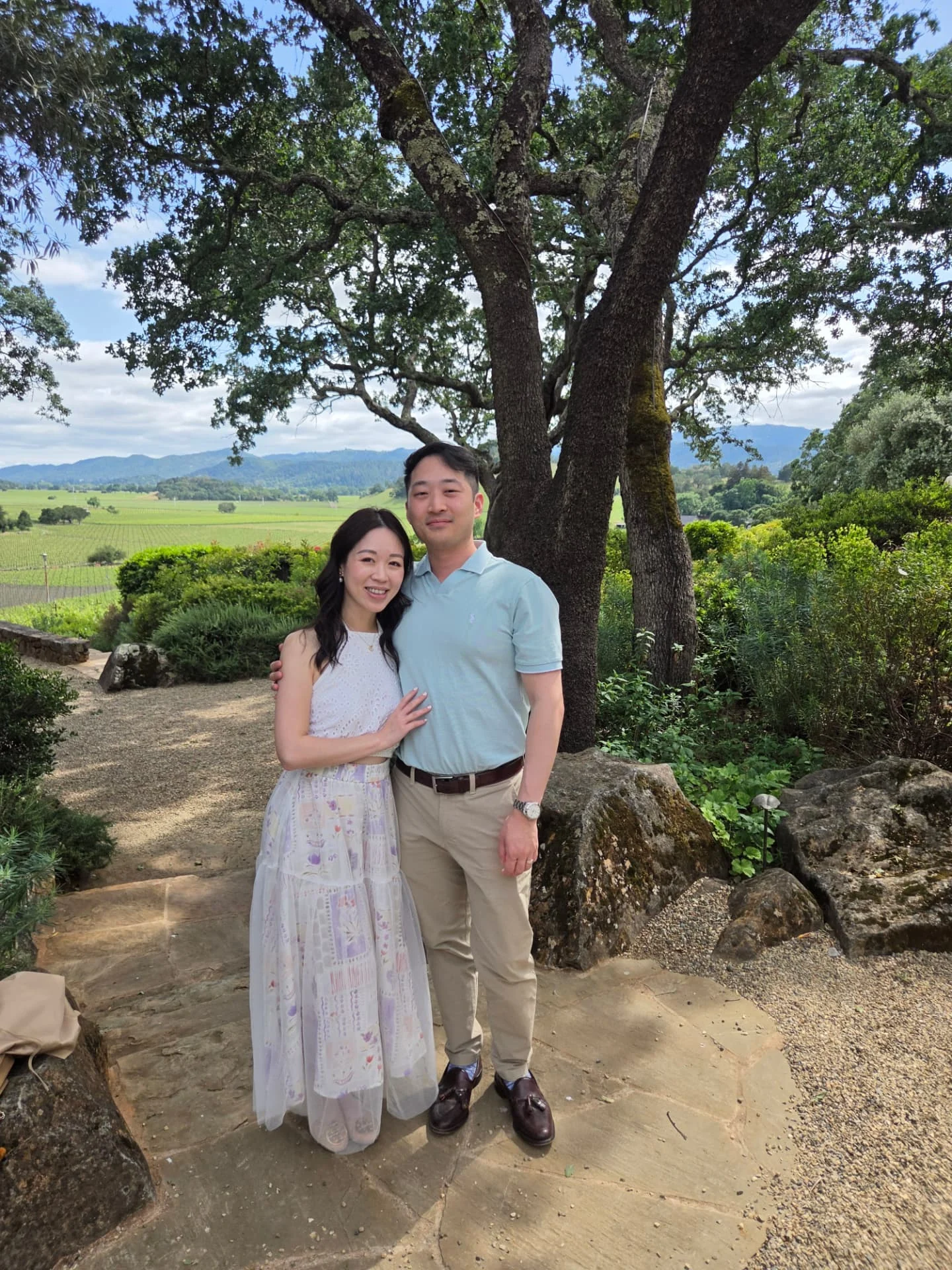 A couple beneath a large tree overlooking vineyards and hills.